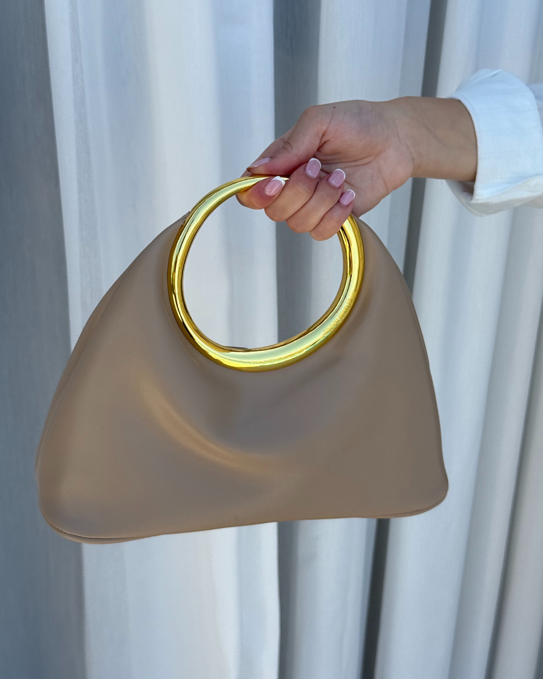 Zena drzi elegantnu beige torbu sa zlatnom ruckom.