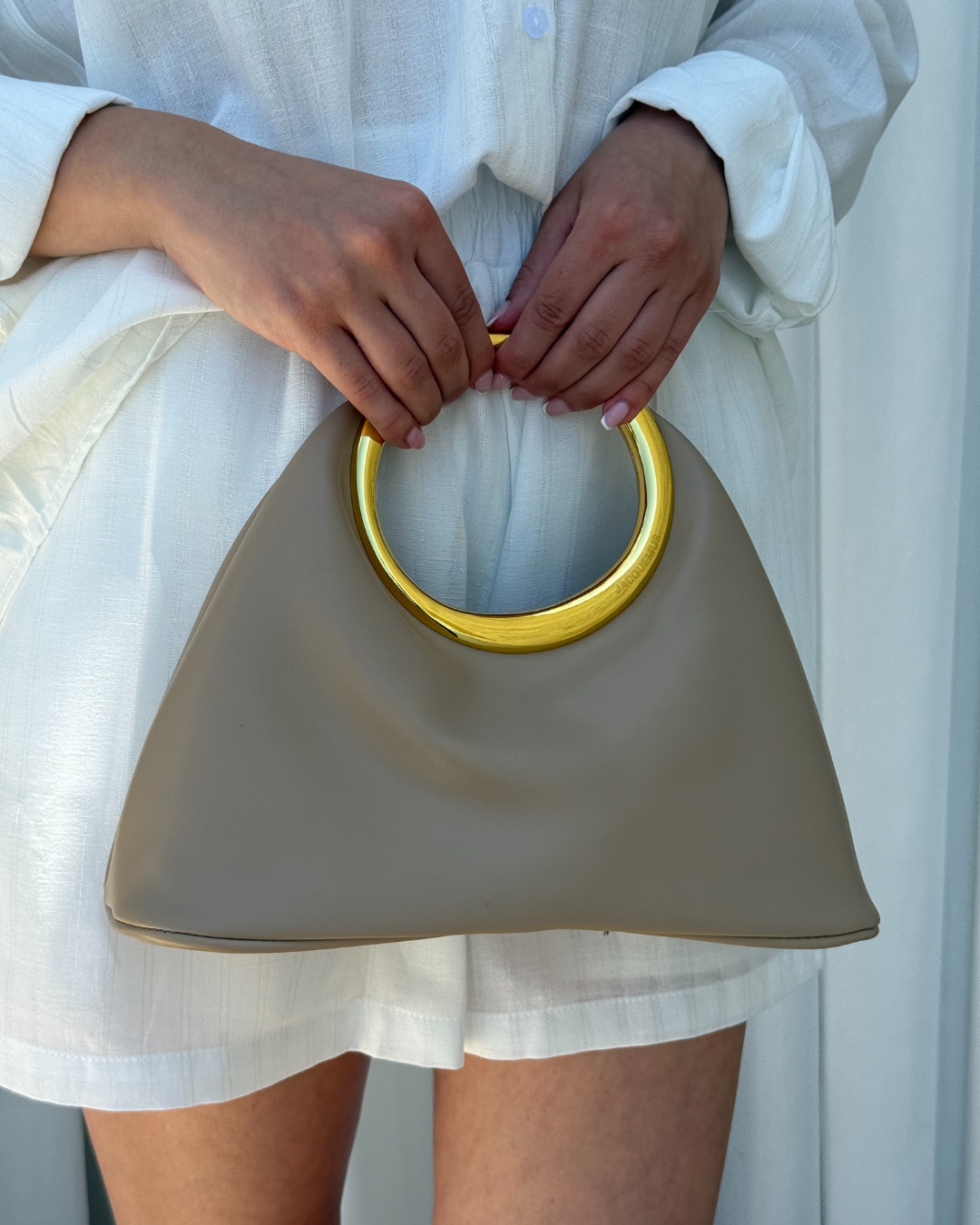 Zena drzi elegantnu beige torbu sa zlatnom ruckom.